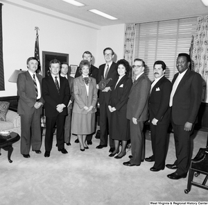 ["Senator John D. (Jay) Rockefeller holds a West Virginia-shaped wall clock and stands among a large group of unidentified visitors in his office."]%