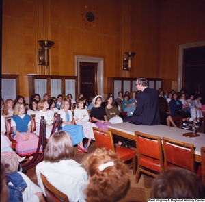 ["This color photograph shows Senator John D. (Jay) Rockefeller sitting on the corner of a table and speaking to a large group of female students."]%