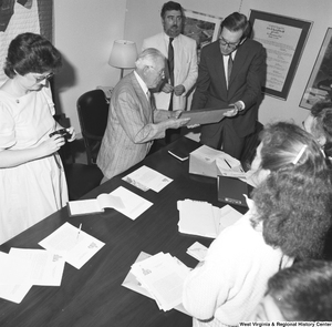 ["Senator John D. (Jay) Rockefeller stands around a conference table and holds a photo that one of the unidentified individuals has given him."]%