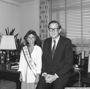 ["Senator John D. (Jay) Rockefeller sits on the corner of the desk in his office and poses for a photograph with a West Virginia woman who wears a pageant sash."]%