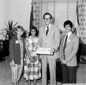 ["Three unidentified individuals with Presidential Classroom for Young Americans badges pose for a photograph with Senator John D. (Jay) Rockefeller in his office as he holds a cake that reads \"Happy Birthday West Virginia: 1863-1985\"."]%