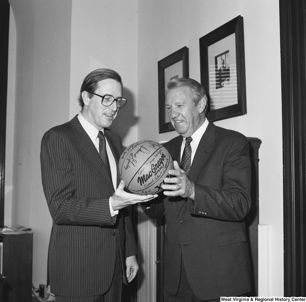["Senator John D. (Jay) Rockefeller holds a basketball with an unidentified man in his office."]%