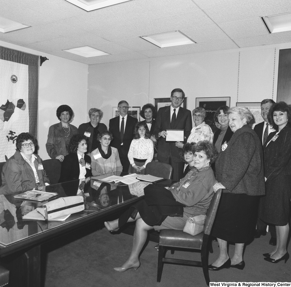 ["Senator John D. (Jay) Rockefeller holds a plaque with members of a large unidentified group in his office."]%