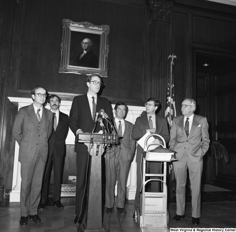 ["Senator John D. (Jay) Rockefeller speaks behind a podium in one of the Senate buildings during a press conference for the Staggers Rail Reform bill. West Virginia Congressmen Nick Rahall and Bob Wise can be seen standing behind him."]%