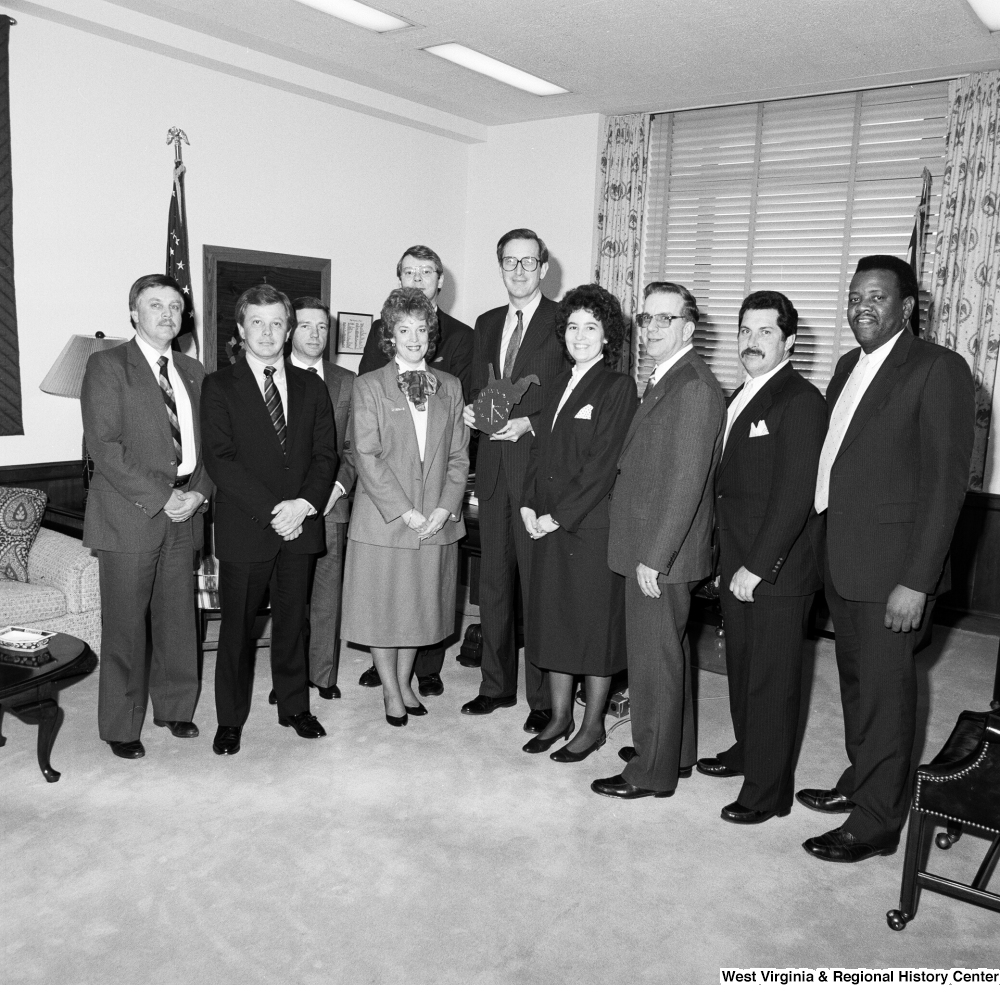 ["Senator John D. (Jay) Rockefeller holds a West Virginia-shaped wall clock and stands among a large group of unidentified visitors in his office."]%