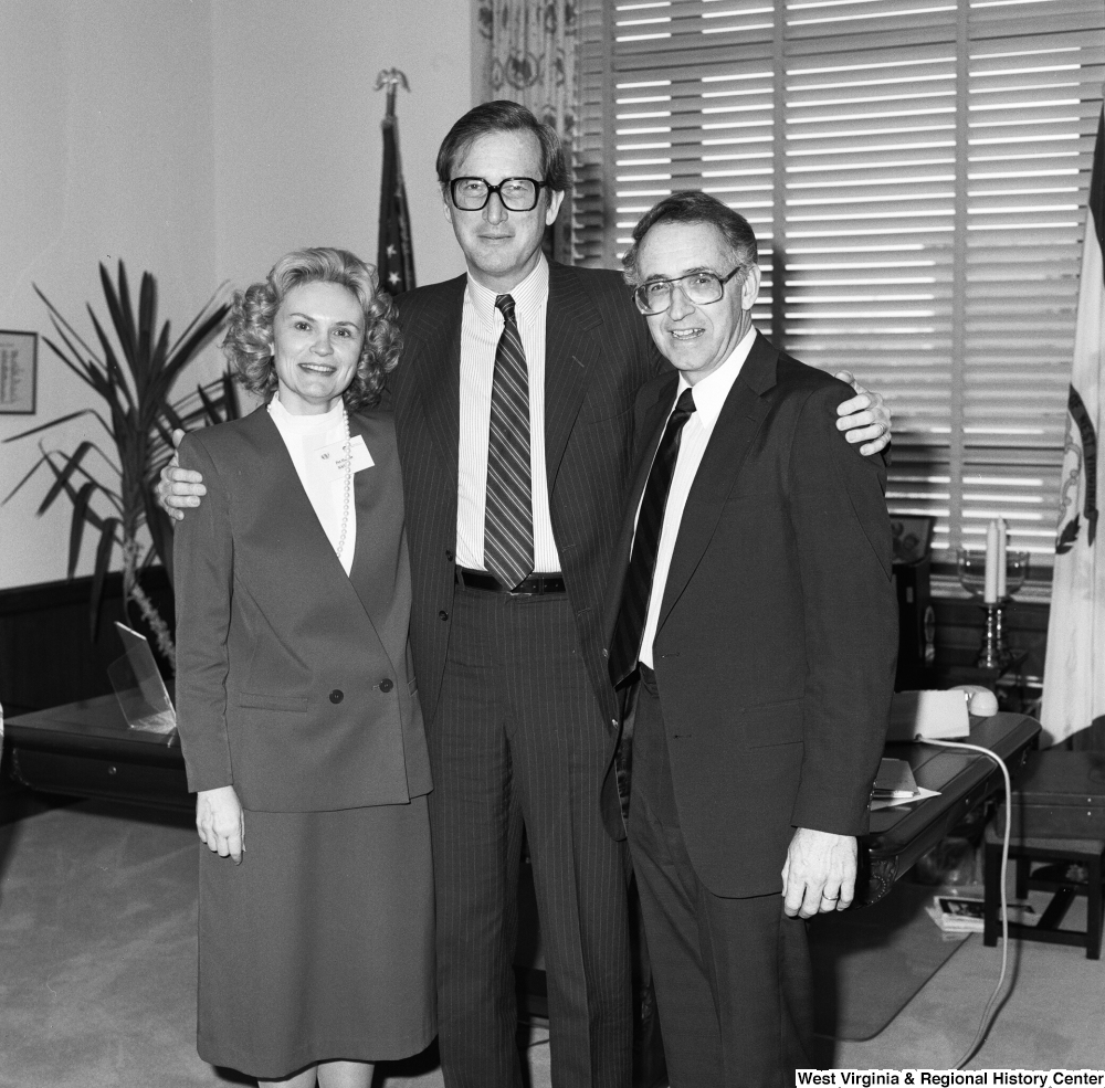 ["Senator John D. (Jay) Rockefeller poses for a photograph with and embraces two unidentified guests in front of the desk in his Washington office. The woman in the photograph has a name badge on that appears to say NASA on it."]%