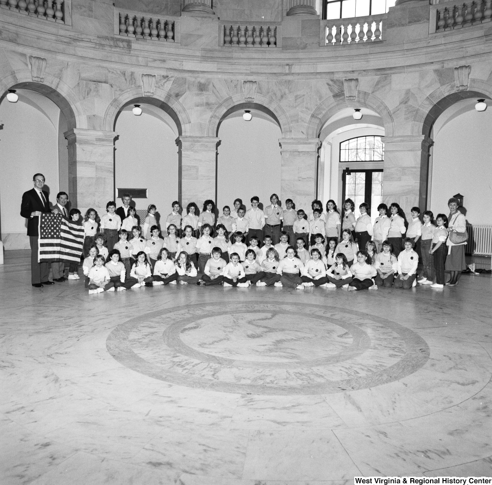 ["Senator John D. (Jay) Rockefeller and Congressman Nick Rahall hold an american flag and pose for a photograph with a large group of young students in a rotunda."]%