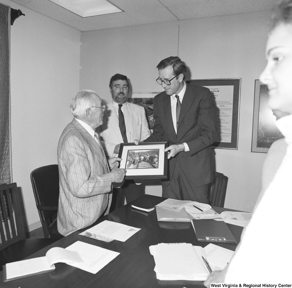["Senator John D. (Jay) Rockefeller holds a photograph of people working on a railroad with an unidentified man."]%