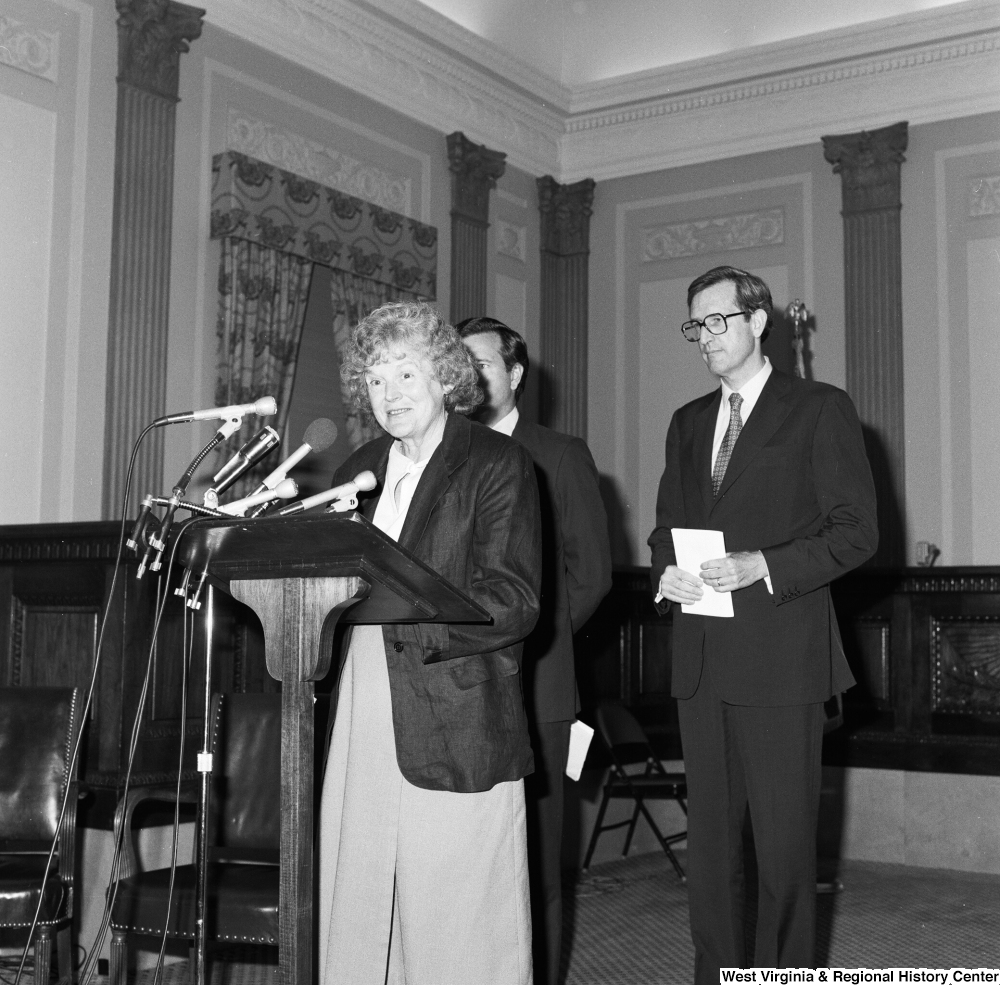 ["Senator John D. (Jay) Rockefeller and Senator John Heinz stand behind a podium where an unidentified woman speaks about the Dislocated Workers Improvement Act."]%