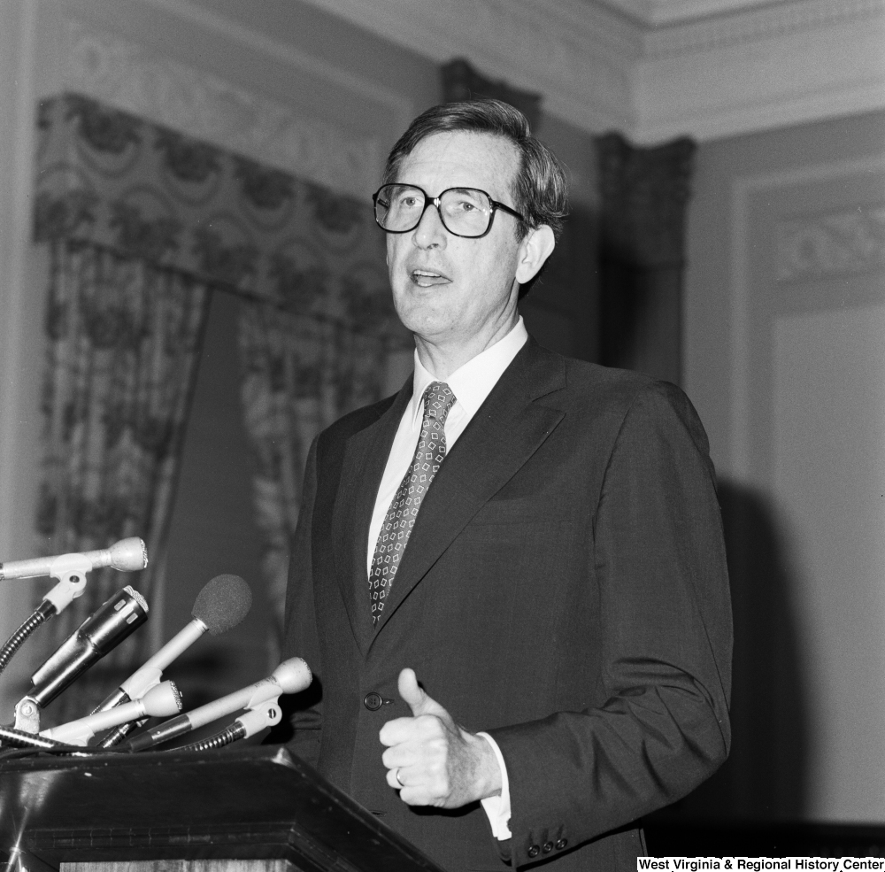 ["Raising a thumb, Senator John D. (Jay) Rockefeller speaks from behind a podium in a Senate building."]%