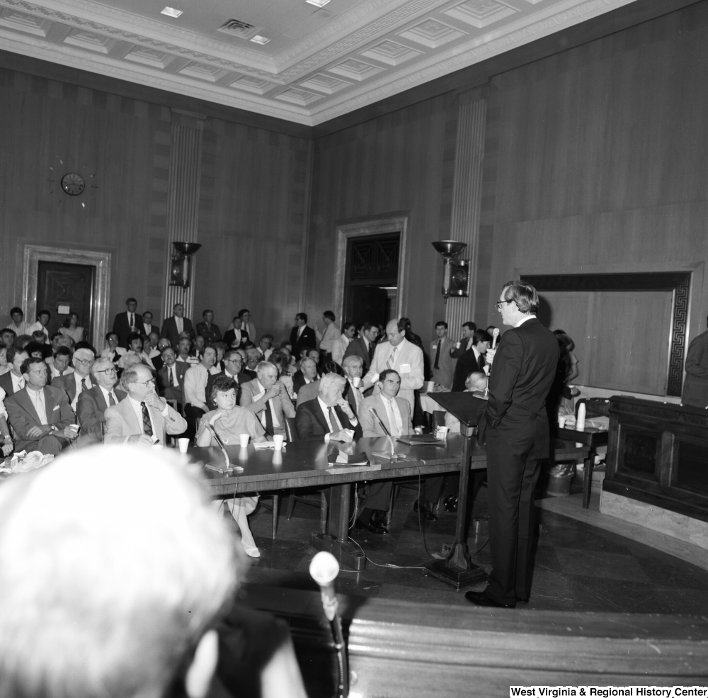 ["Senator John D. (Jay) Rockefeller addresses a large audience in one of the Senate committee hearing rooms."]%