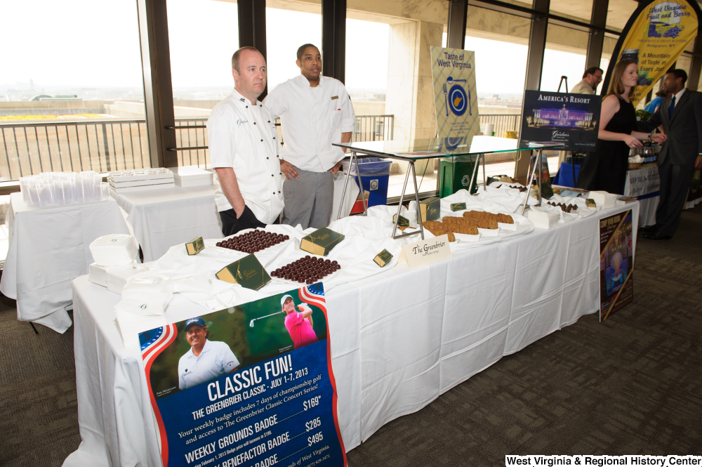 ["Two men from the Greenbrier stand behind their table at the 150th birthday celebration of West Virginia."]%