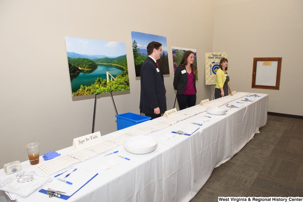 ["Three people stand behind the sign in table for the 150th birthday celebration for West Virginia."]%