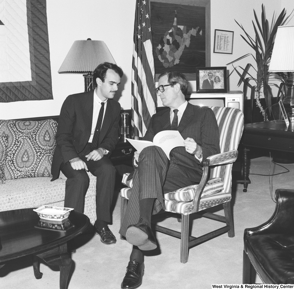 ["Senator John D. (Jay) Rockefeller flips through a document and speaks with an unidentified man in his office."]%