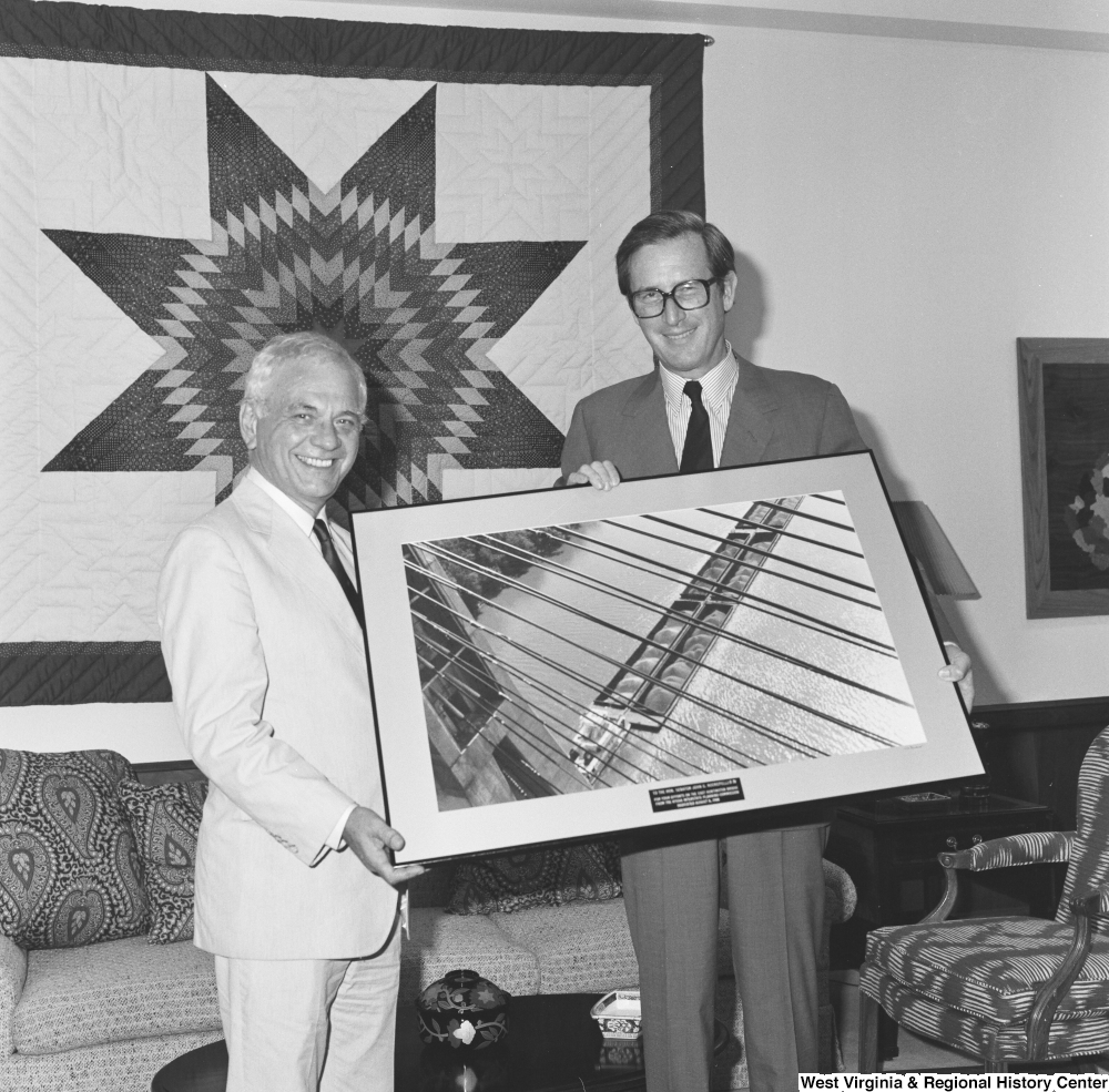 ["Senator John D. (Jay) Rockefeller holds a framded photograph of a coal barge crossing under a bridge, an apparent gift from the unidentified man on his right."]%