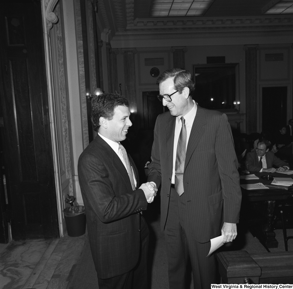 ["Senator John D. (Jay) Rockefeller shakes hands and laughs with an unidentified man in a Senate hearing room."]%