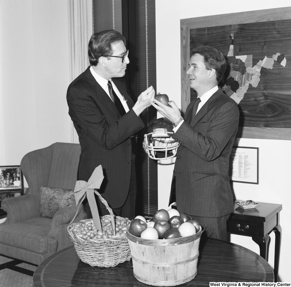 ["Senator John D. (Jay) Rockefeller stands next to Senator John Breaux who is holding an apple and a football helmet."]%